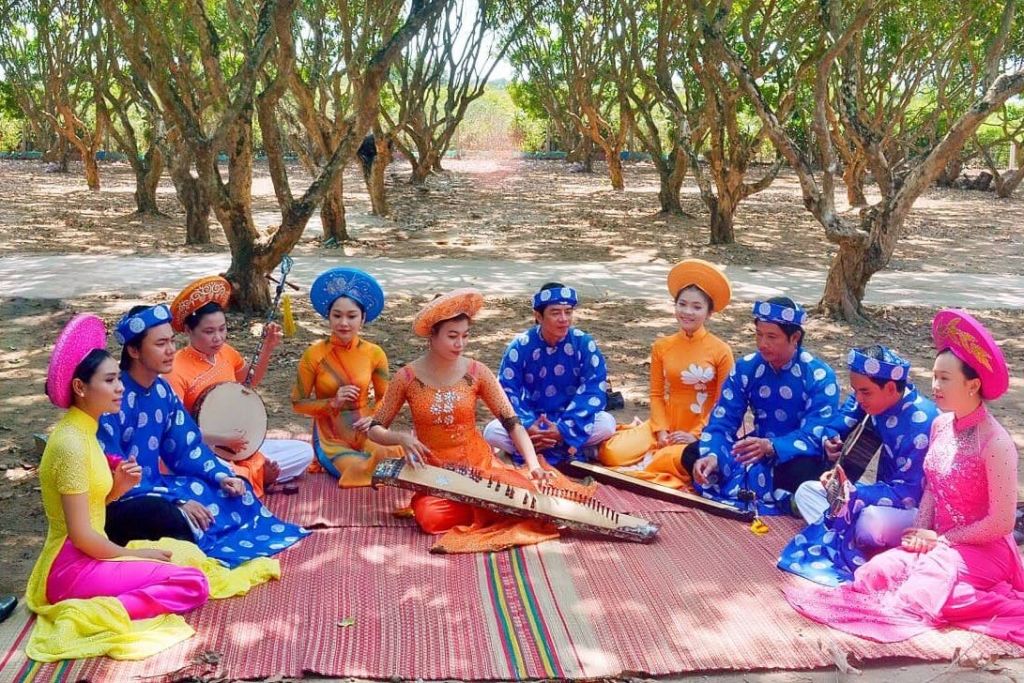 화려한 전통 의상을 입은 사람들이 야외에서 나무 아래 매트에 앉아 악기를 연주합니다.