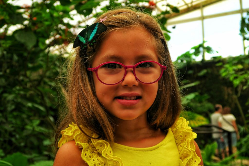 나뭇잎이 무성한 야외 환경에서 노란색 상의를 입고 미소를 지으며 머리카락에 안경을 쓰고 나비를 입은 어린 소녀.