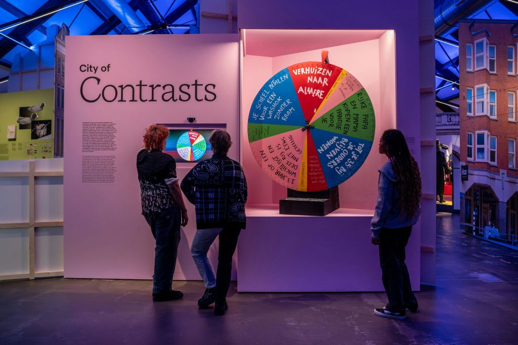 조명이 밝고 현대적인 박물관 공간에 세워진 문자가 담긴 다채로운 휠이 있는 전시관 근처에 세 사람이 서 있습니다.