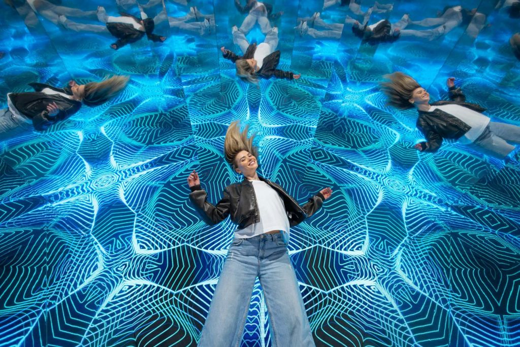 한 여성이 기하학적 파란색 디자인의 패턴 반사 표면에 누워 거울 반사로 둘러싸여 있습니다.