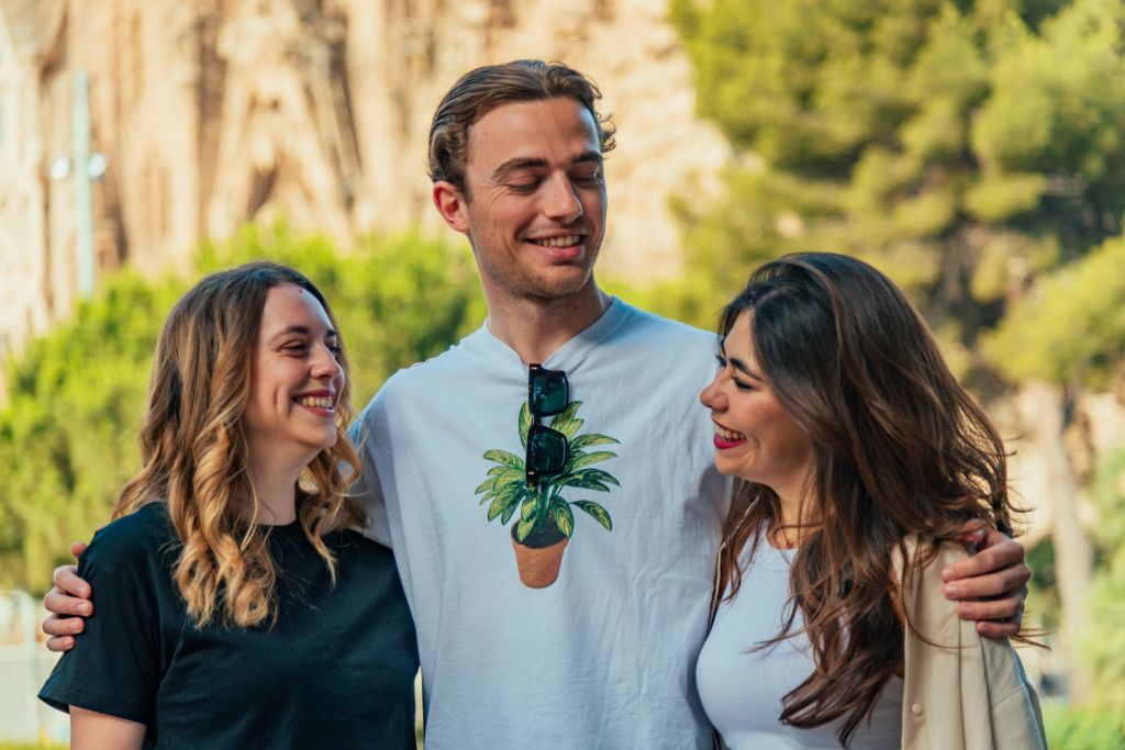 흐릿한 녹지와 구조물을 배경으로 세 사람이 야외에 서서 미소를 짓고 서로를 바라보고 있습니다.