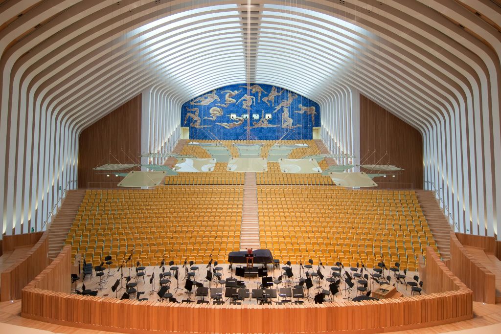 노란색 좌석이 줄지어 있는 텅 빈 오케스트라 콘서트 홀, 무대 위의 그랜드 피아노, 좌석 공간 위의 예술적 벽화.