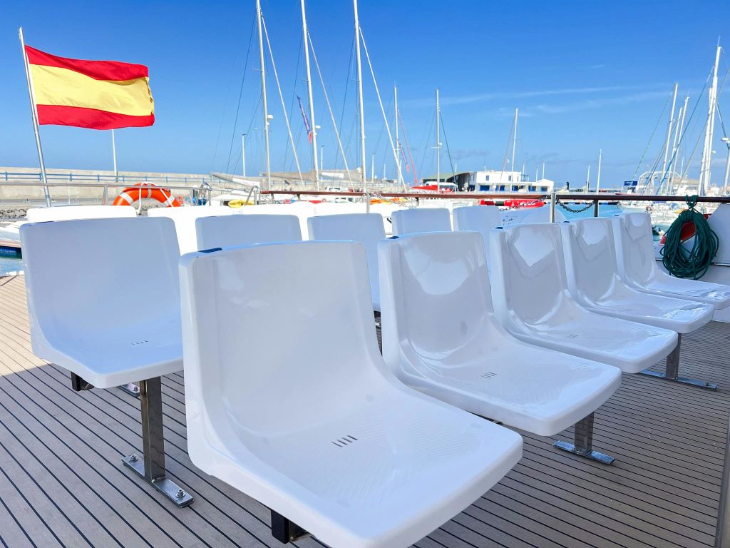 스페인 국기가 달린 보트 갑판의 흰색 플라스틱 좌석과 맑고 푸른 하늘 아래 배경으로 선착장이 딸린 요트 몇 척.