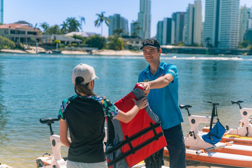 한 남성이 건물을 배경으로 물가 근처에 있는 사람에게 빨간색 구명조끼를 건네고 있습니다.