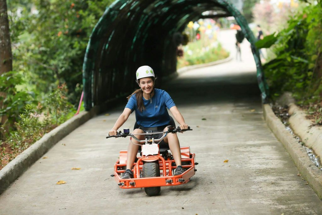 헬멧을 쓴 웃는 사람이 녹음이 우거진 아치 아래 포장된 길에서 주황색 고카트를 타고 있습니다.