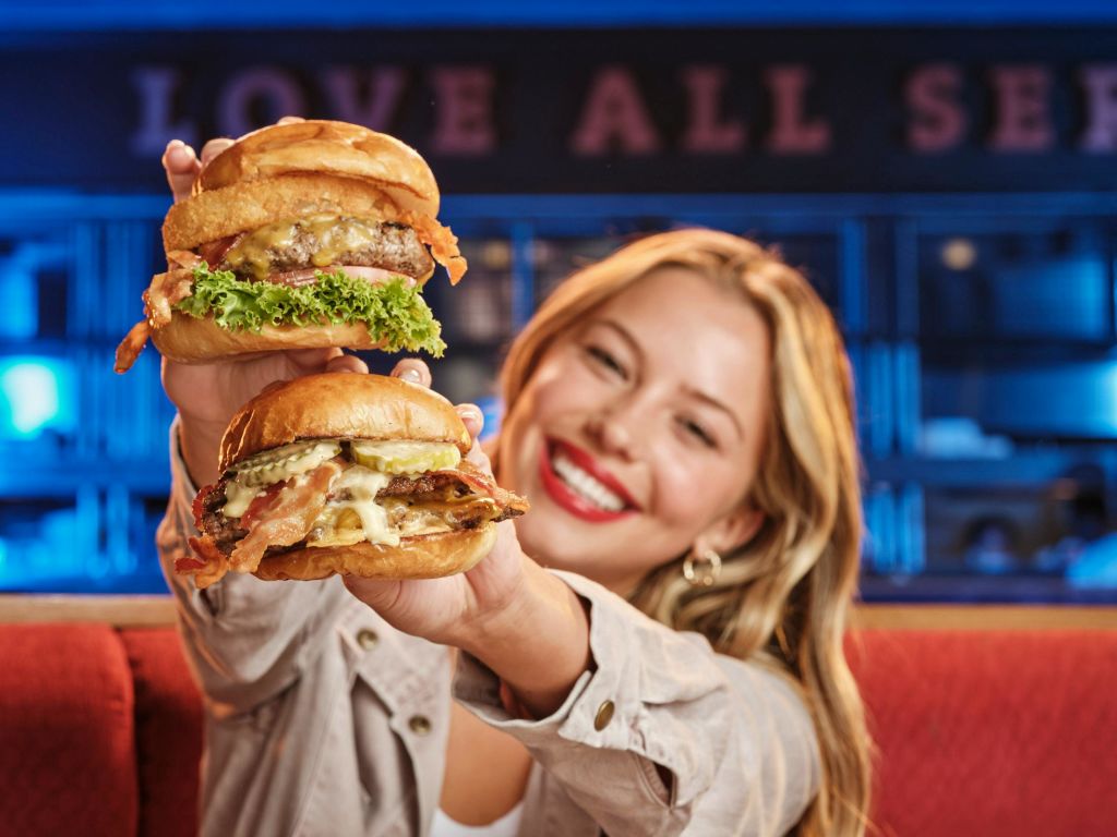 금발에 빨간 립스틱을 바른 여성이 양상추와 베이컨을 곁들인 치즈버거 두 개를 들고 미소를 짓고 있습니다.