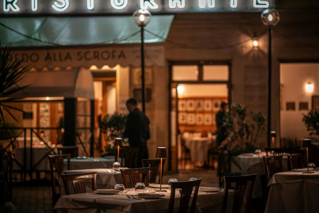 세트 테이블과 유리 제품, 배경의 두 사람, 부분적으로 보이는 조명 표지판이 있는 조명이 희미한 야외 레스토랑입니다.