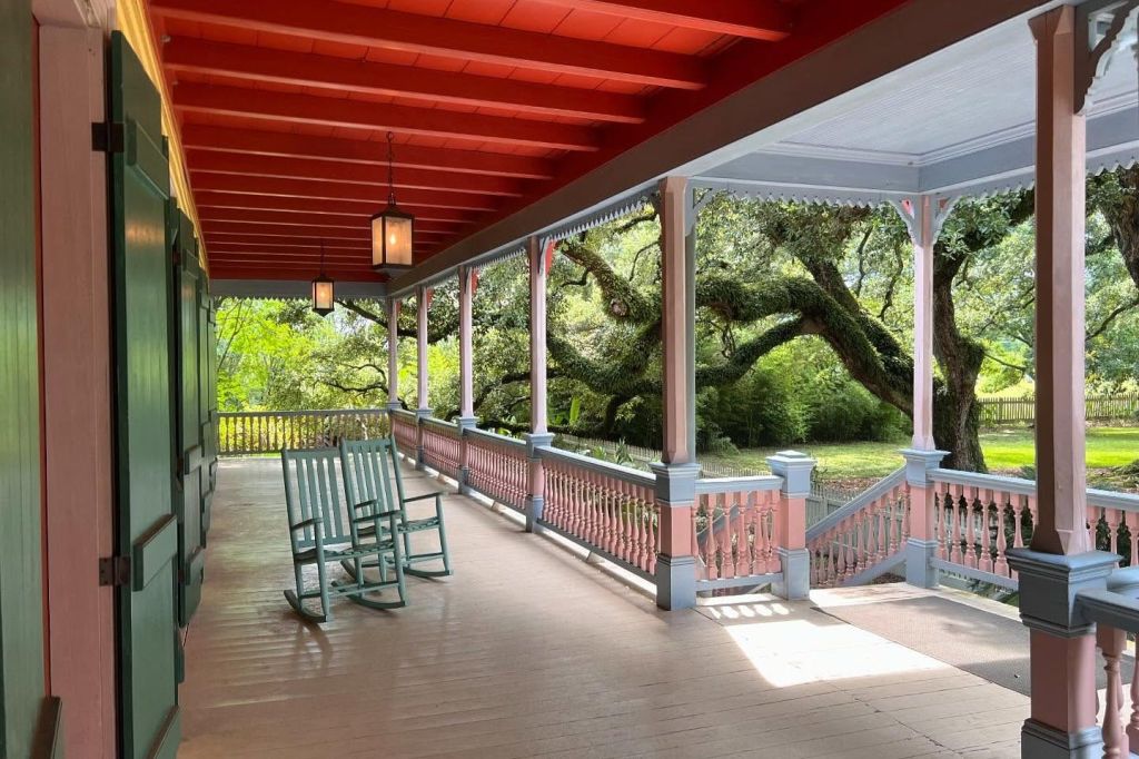 청록색 흔들 의자가 있는 베란다에서는 커다랗고 갈라진 나무가 있는 정원이 내려다보입니다. 베란다에는 빨간색 천장과 기둥이 있습니다.