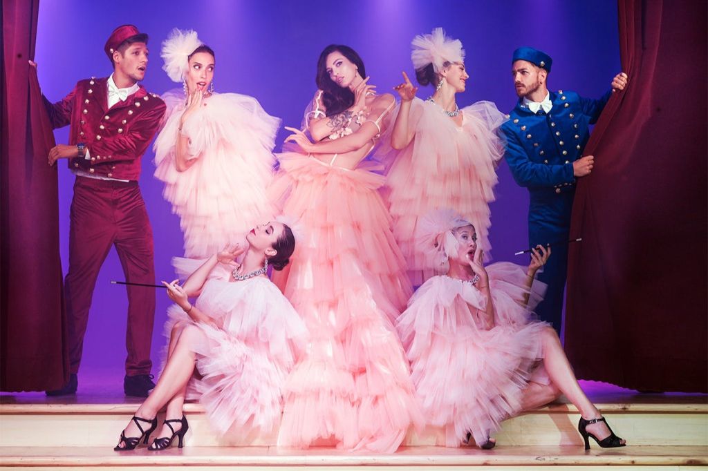 우아한 분홍색 드레스를 입은 공연자와 빨간색 유니폼을 입은 남성이 보라색 배경의 무대에 서 있습니다.