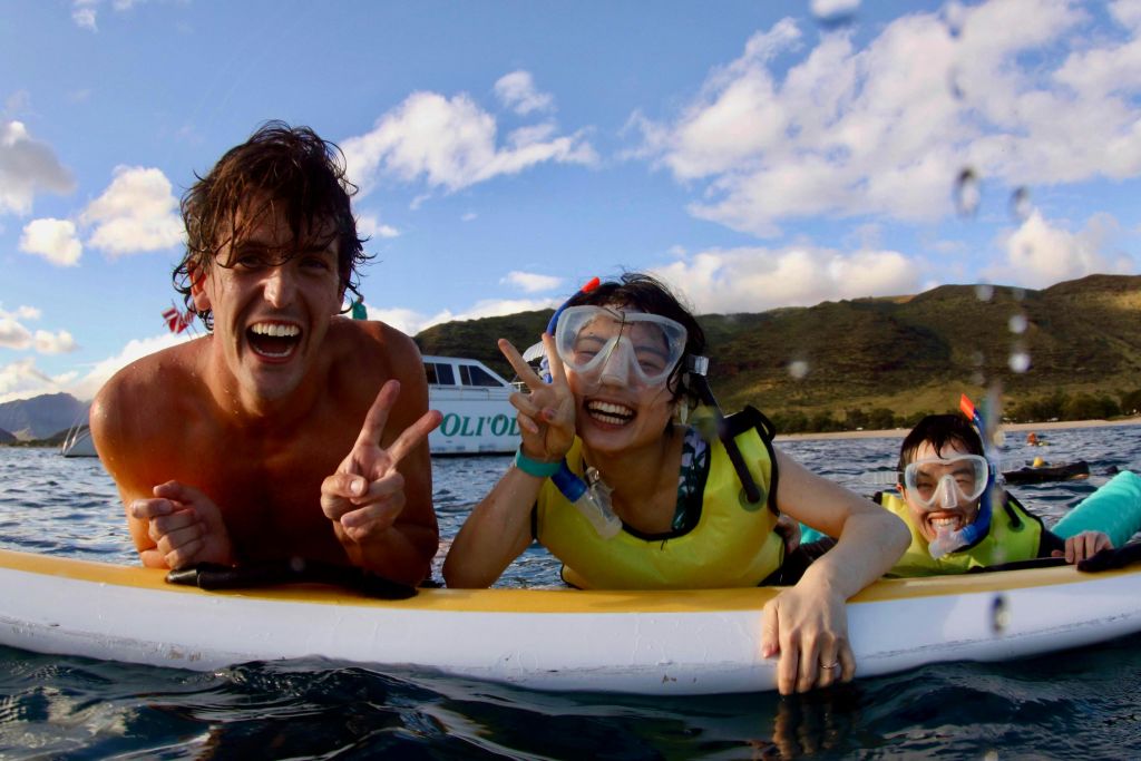 바다에 떠 있는 노란색 보드에 스노클링 장비를 착용한 두 사람이 미소를 짓고 평화 표지판을 반짝이며 보트와 산을 배경으로 사진을 찍고 있습니다.