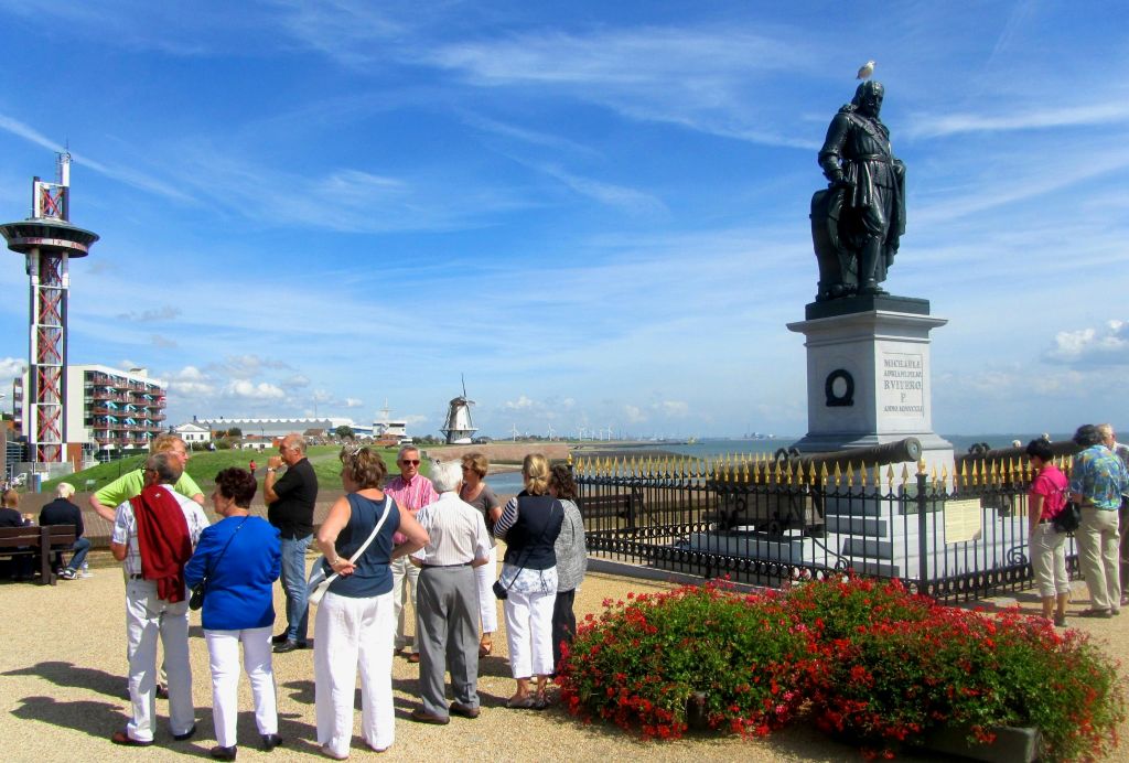 한 무리의 사람들이 울타리로 둘러싸인 새가 꼭대기에 있는 동상 근처에 서 있습니다. 풍차와 해안선이 배경으로 보입니다.