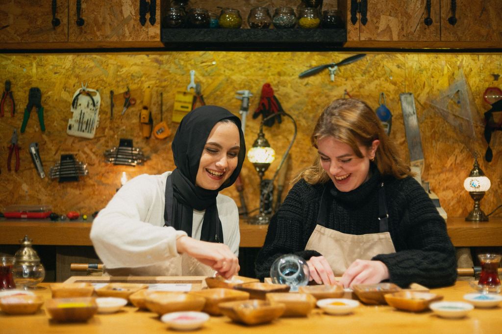 도구로 둘러싸인 공방에서 모자이크 램프를 켜고 미소를 짓는 두 여성.