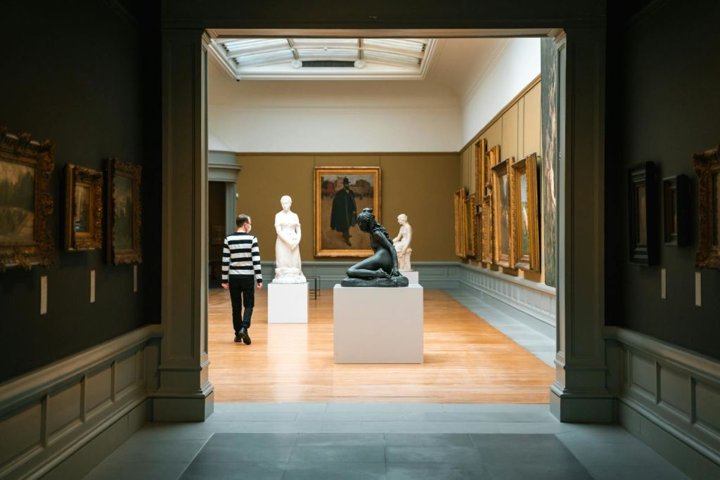 줄무늬 셔츠를 입은 사람이 조각품, 액자 그림, 나무 바닥이 있는 미술관을 거닐고 있습니다.