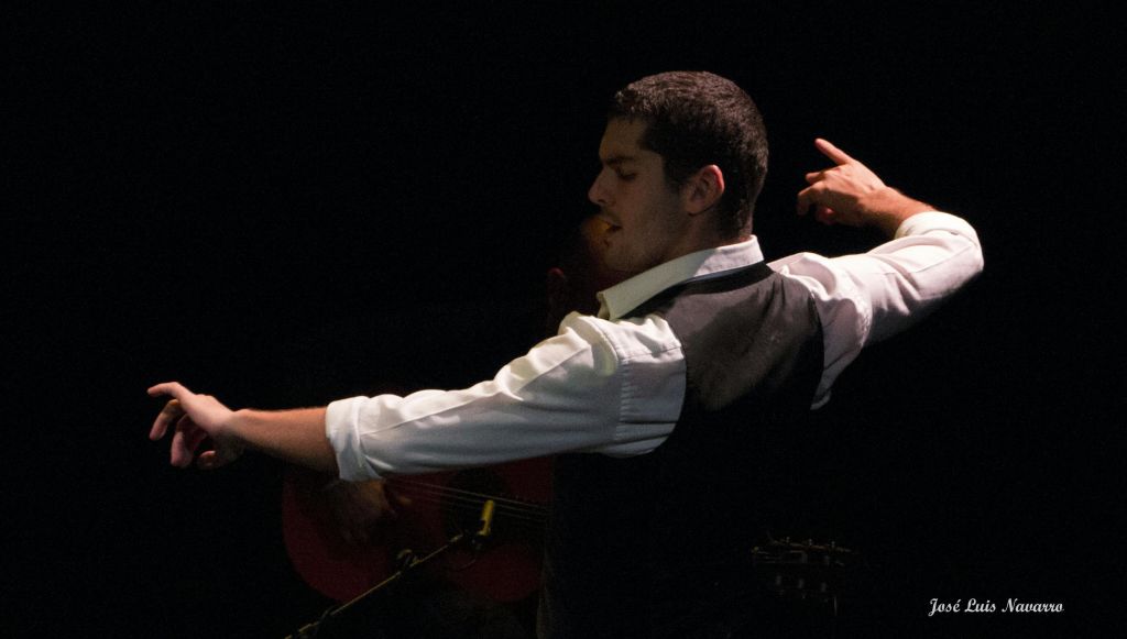 부분적으로 보이는 기타와 뮤지션이 있는 어두운 배경에 흰색 셔츠와 어두운 조끼를 입고 춤을 추는 남성.