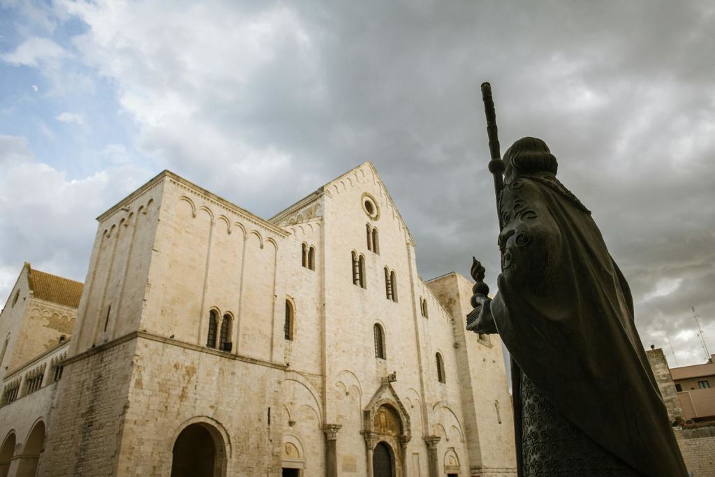 지팡이를 들고 있는 조각상이 흐린 하늘 아래 크고 유서 깊은 석조 건물 앞에 서 있습니다.
