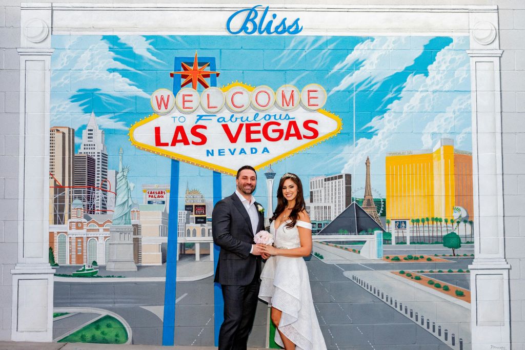 웰컴 투 라스베이거스 (Welcome to Las Vegas) 표지판의 화려한 벽화 앞에 한 커플이 서 있으며, 도시 랜드마크와 그 위에 'Bliss' 라는 단어가 있습니다.