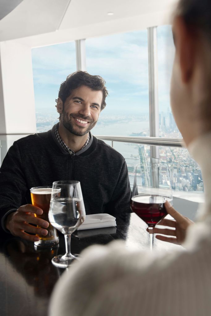 한 남자가 레드 와인 한 잔을 들고 누군가의 맞은편에서 맥주를 들고 미소를 짓고 있습니다. 도시 전망을 감상할 수 있는 창문 근처에 자리잡고 있습니다.