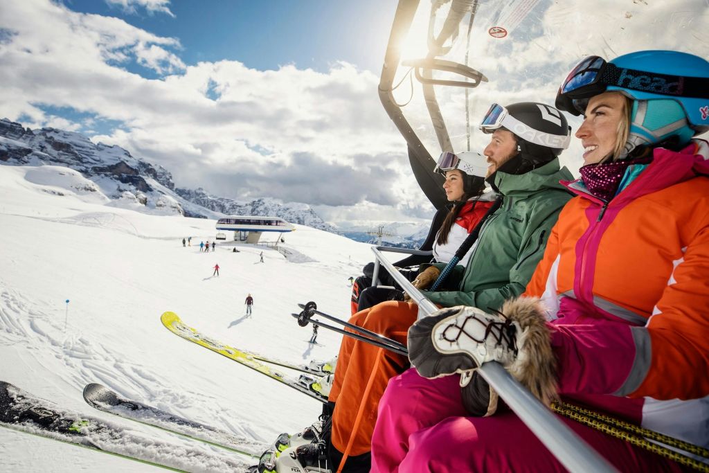 눈 덮인 산과 스키어를 배경으로 화창한 날씨에 스키 리프트를 타는 겨울 장비를 입은 세 사람.