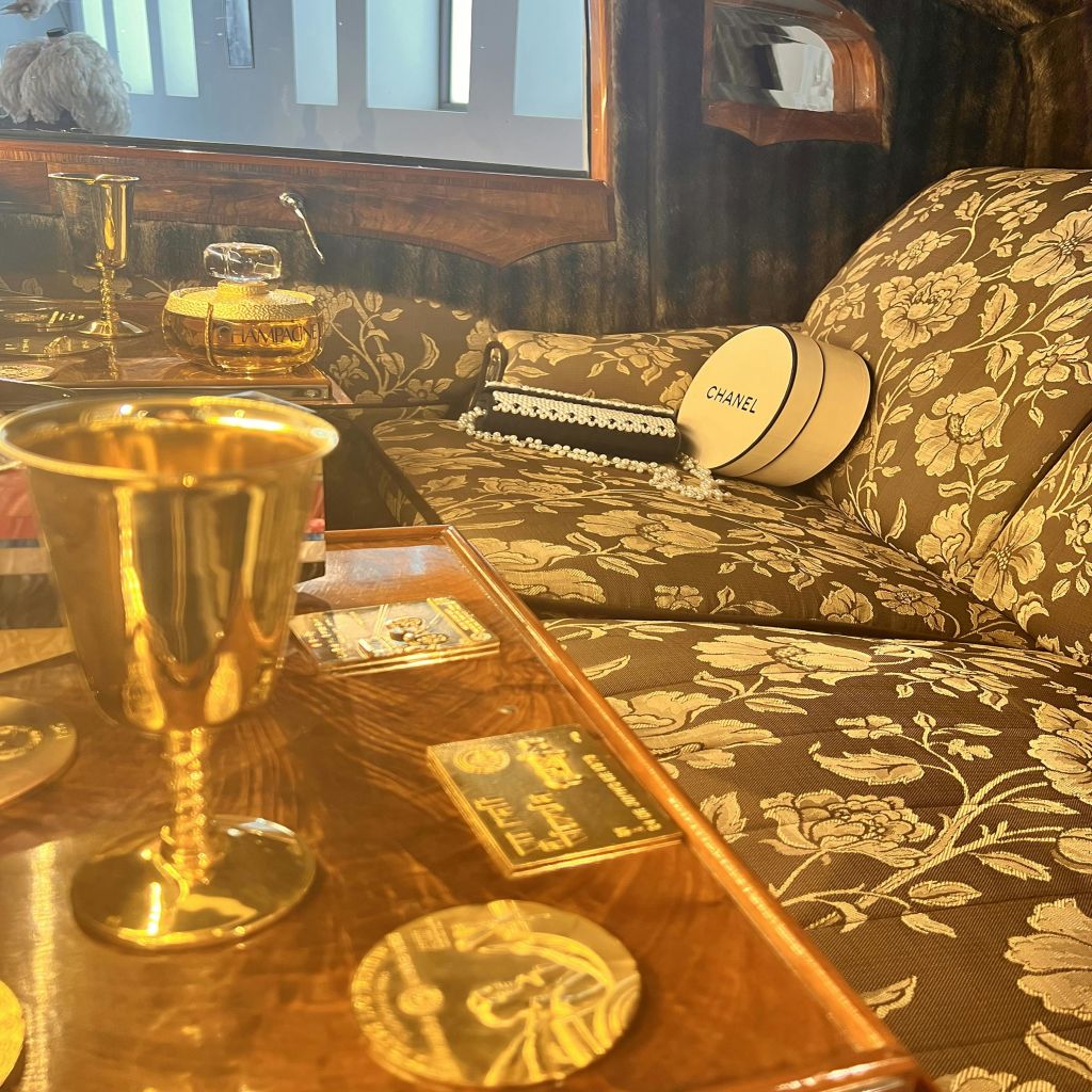 꽃무늬 의자, 나무 테이블에 놓인 금잔, 소파에 놓인 샤넬 모자 상자가 있는 고급스러운 인테리어.