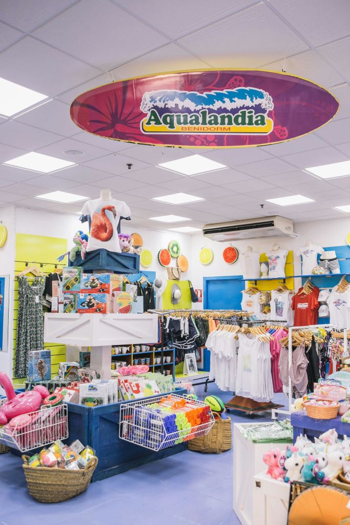 옷, 장난감, 기념품 등 다채로운 상품이 있는 기프트숍 인테리어. "Aqualandia Benidorm" 표지판이 머리 위에 걸려 있습니다.