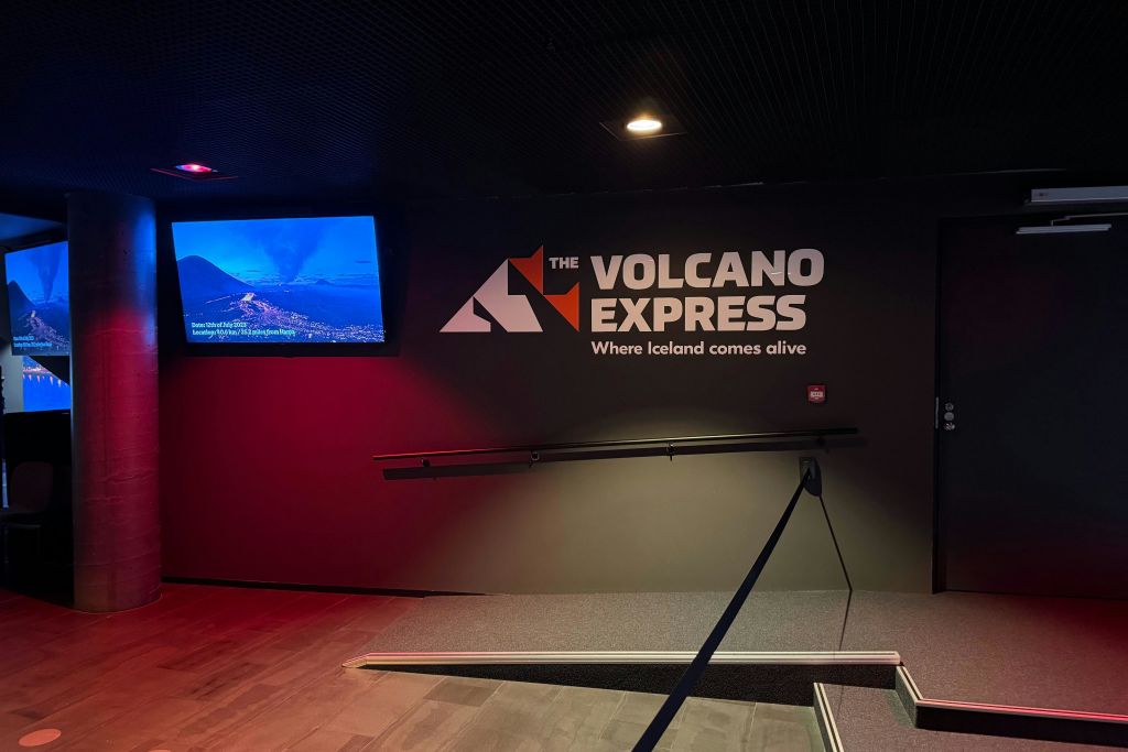 "The Volcano Express: Where Iceland comes alive (화산 특급: 아이슬란드가 살아 숨쉬는 곳)" 이라는 조명 표지판과 풍경 이미지를 보여주는 화면이 있는 어두운 복도.