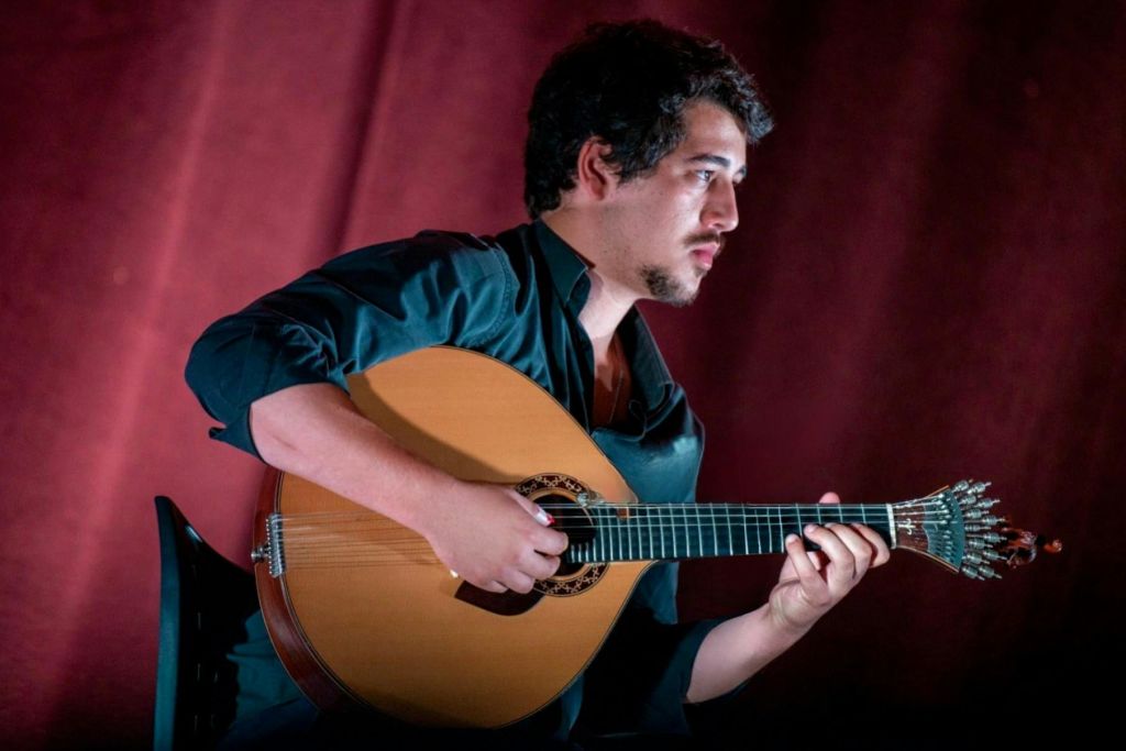 검은색 셔츠를 입고 어두운 빨간색 배경으로 현악기를 연주하는 검은색 머리카락과 수염을 가진 사람.