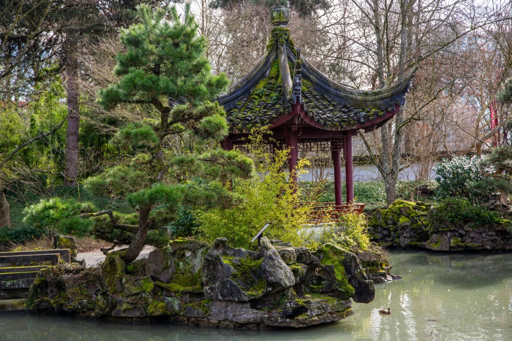 고요한 정원의 바위, 나무, 녹지로 둘러싸인 연못 옆에 곡선형 지붕의 전통 파빌리온이 있습니다.