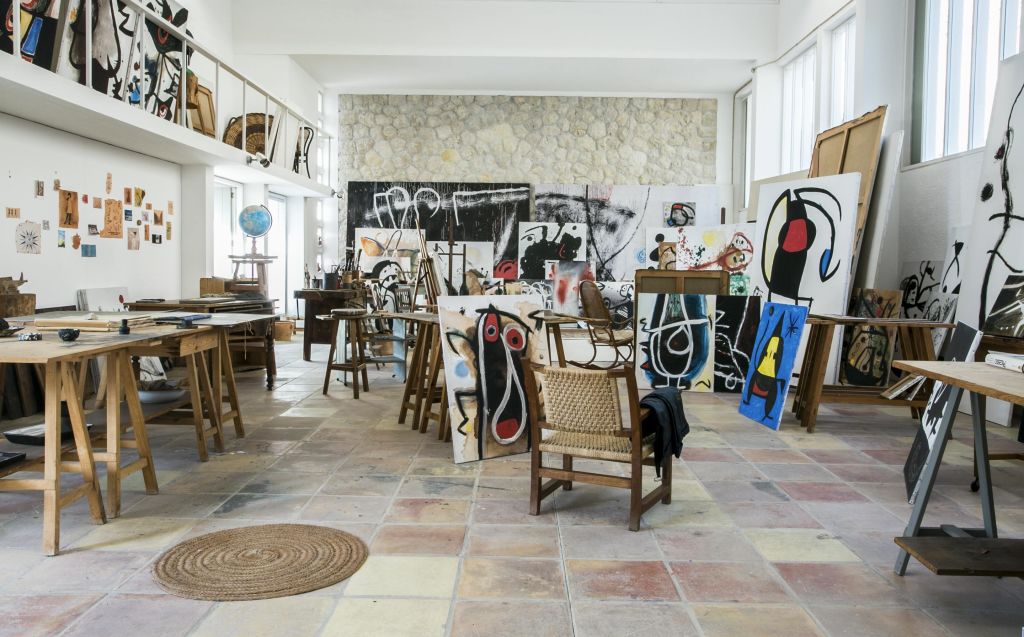 이젤과 바닥에 추상적인 그림, 나무 의자, 테이블, 지구본이 있는 아트 스튜디오. 배경에는 돌담과 타일 바닥이 있습니다.