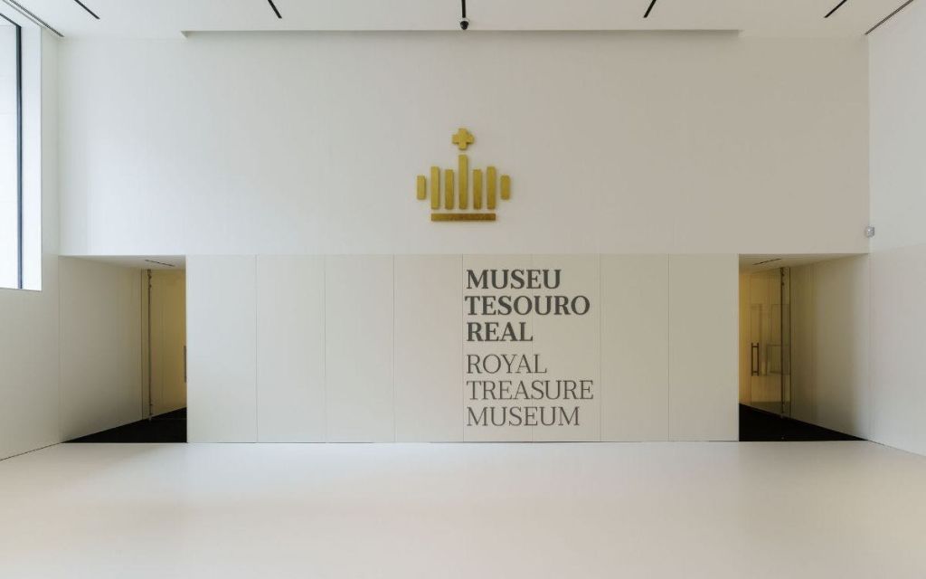 황금 왕관 기호 아래에 "Museu Tesouro Real" 과 "Royal Treasure Museum" 텍스트가 있는 벽. 화이트와 미니멀한 디자인.