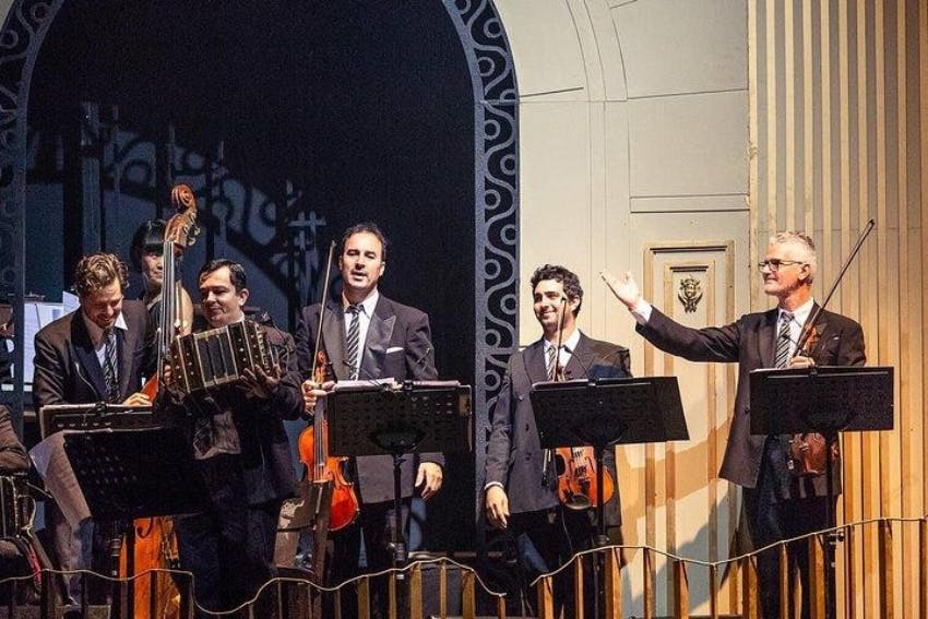 수트를 입은 5명의 뮤지션이 무대에서 첼로, 아코디언, 바이올린 등 다양한 악기를 들고 어두운 조명 아래에서 공연합니다.