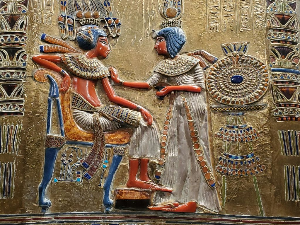 황금색 배경에 손을 얹은 정교한 머리 장식과 보석으로 정교한 복장을 한 두 인물을 묘사한 고대 이집트 구호품.