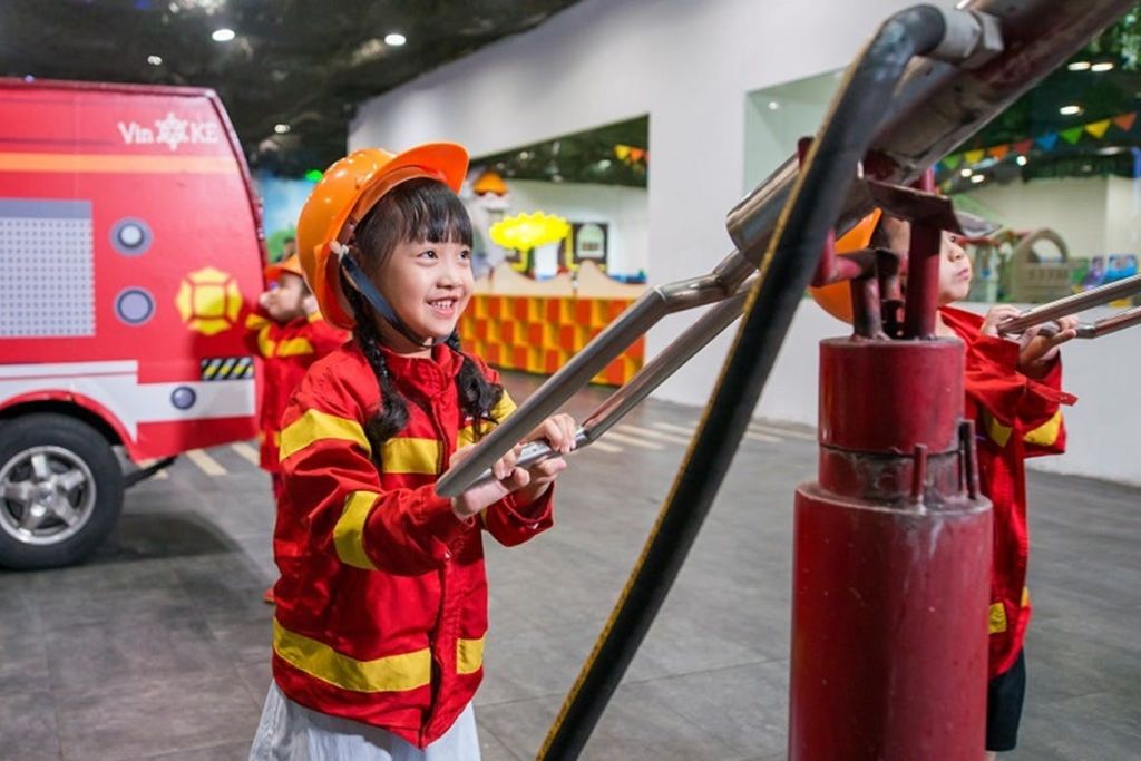 빨간색과 노란색 소방관 유니폼과 헬멧을 착용한 어린 소녀가 실내 놀이 공간의 작은 소방차 근처에서 도구를 사용합니다.