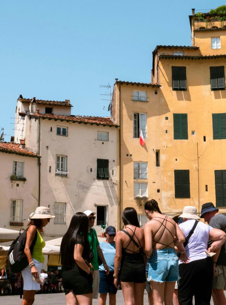 맑고 푸른 하늘 아래 노란색과 흰색 외관이 있는 오래된 건물 앞에 한 무리의 사람들이 여름 옷을 입고 모여듭니다.