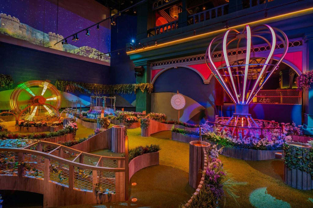 화려한 조명, 곡선형 통로, 식물, 밤하늘 벽화 아래 기발하고 커다란 장식을 갖춘 실내 미니 골프 코스.