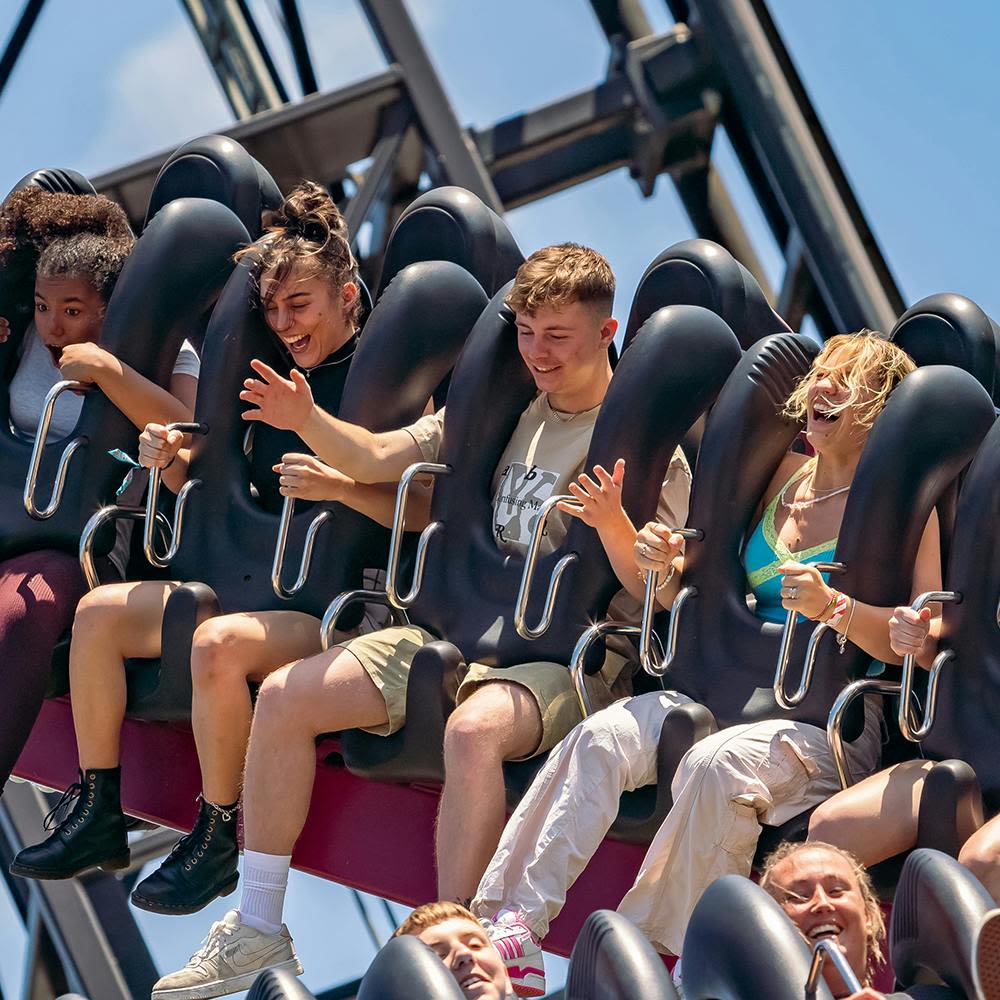 푸른 하늘과 금속 구조물을 배경으로 일시 정지된 롤러코스터를 탄 사람들이 미소를 지으며 구속을 유지하고 있습니다.