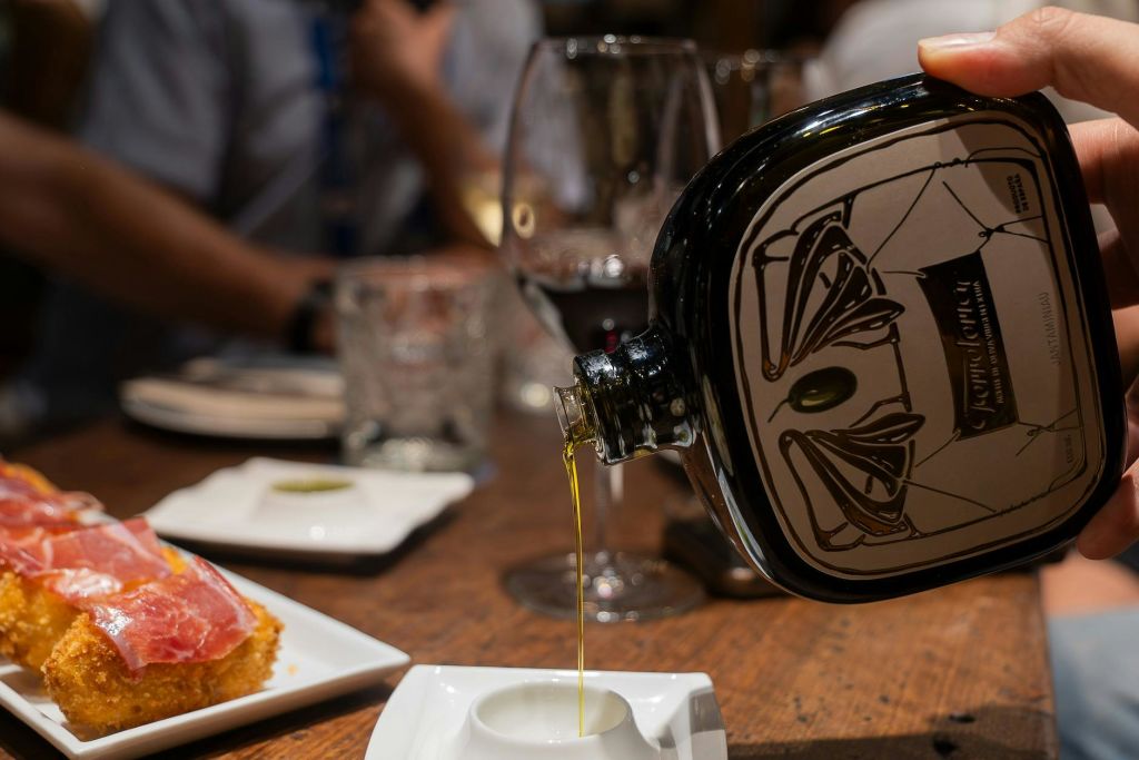 유리병은 유리잔과 접시가 놓인 나무 테이블 위에 놓인 햄에 얹은 애피타이저 접시 옆에 놓인 작은 접시에 기름을 붓습니다.