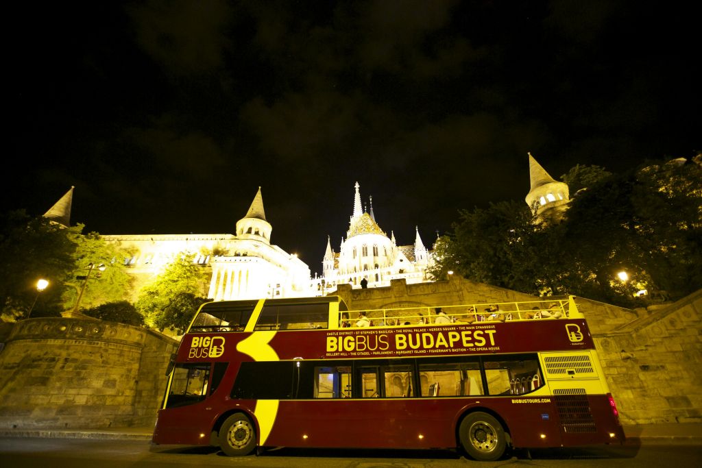 밤에는 '빅 버스 부다페스트' 라는 이름의 빨간색 2층 투어 버스가 조명이 켜진 역사적인 건물을 배경으로 주차되어 있습니다.
