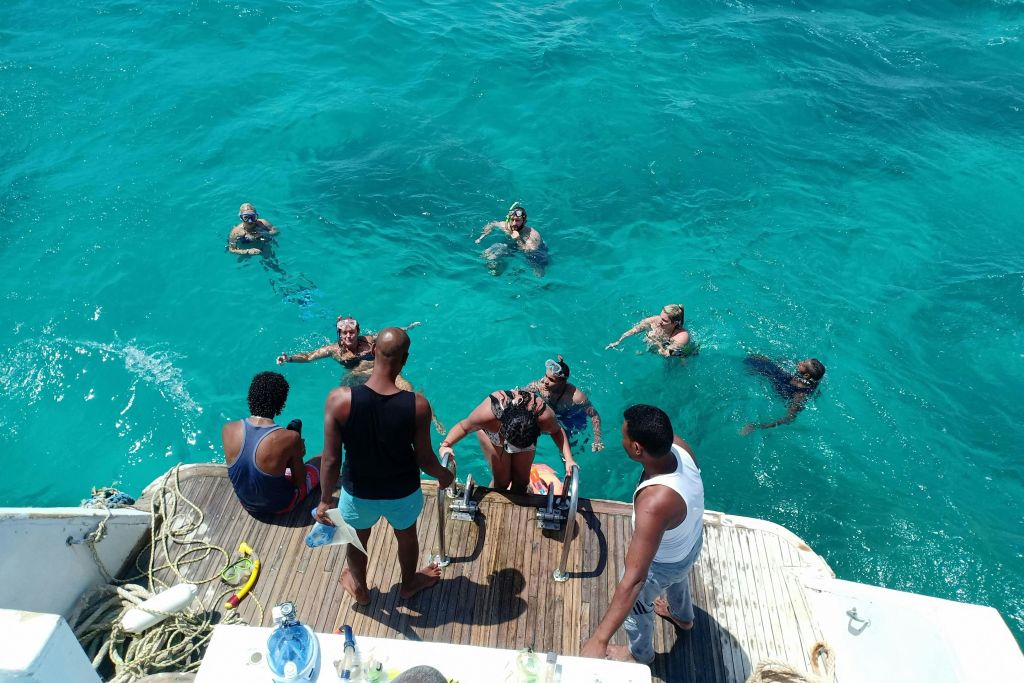 보트 옆의 맑고 푸른 물에서 수영을 즐기는 사람들. 일부는 선상에 있고 다른 일부는 보트 근처의 물에 있습니다.