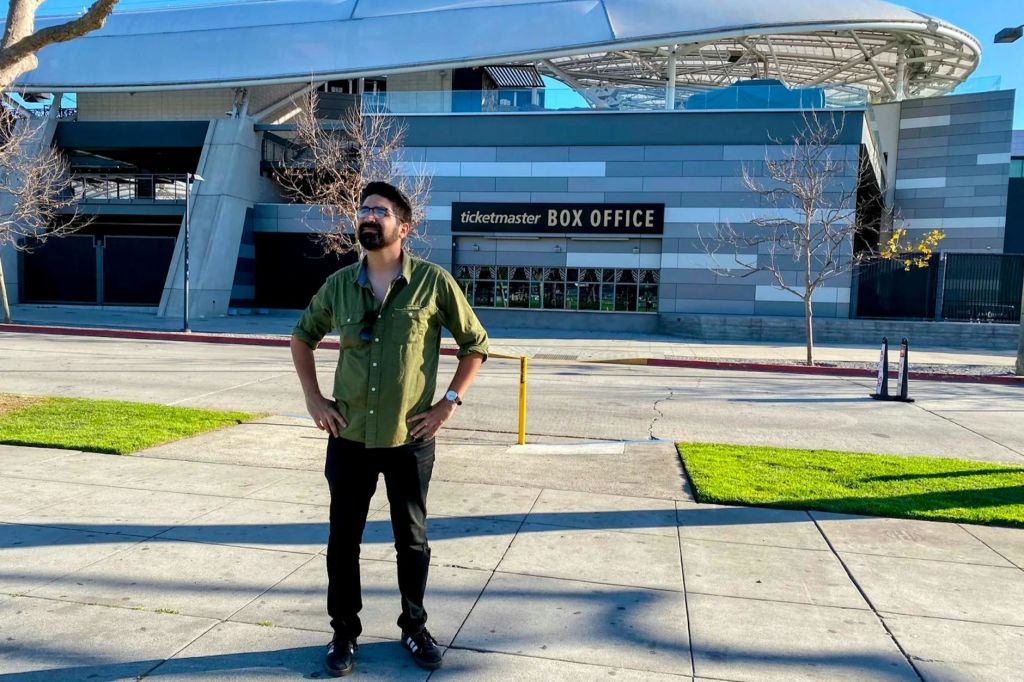 초록색 셔츠를 입은 한 남성이 Ticketmaster 박스 오피스 근처의 보도에 서서 현대적인 건물과 나뭇잎이 없는 나무를 배경으로 서 있습니다.