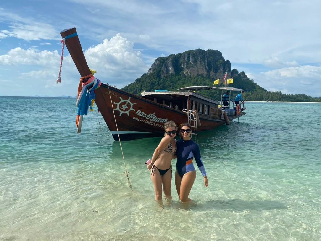 수영복을 입은 두 사람이 산악 섬을 배경으로 보트 근처의 얕은 물에 서 있습니다.