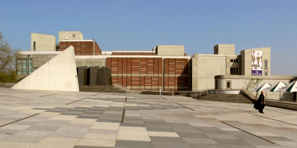 맑고 푸른 하늘 아래 광활한 타일 광장으로 둘러싸인 기하학적 디자인의 대형 벽돌과 콘크리트 건물.