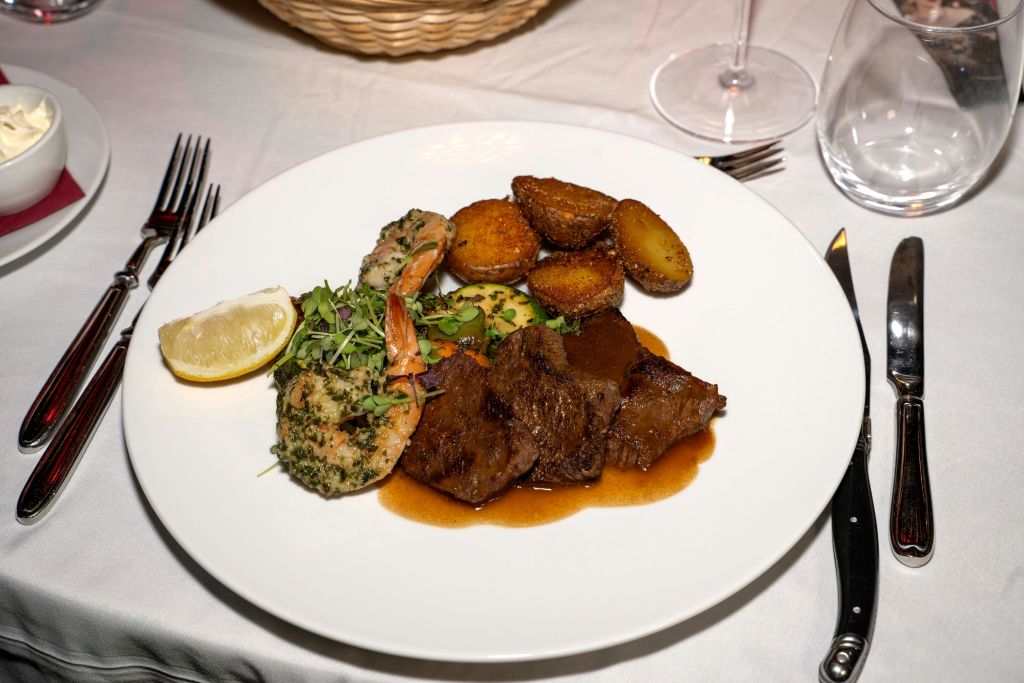 식기류가 담긴 흰색 식탁보에 양념 새우, 스테이크, 구운 감자, 채소, 레몬 웨지를 곁들인 도금 식사.