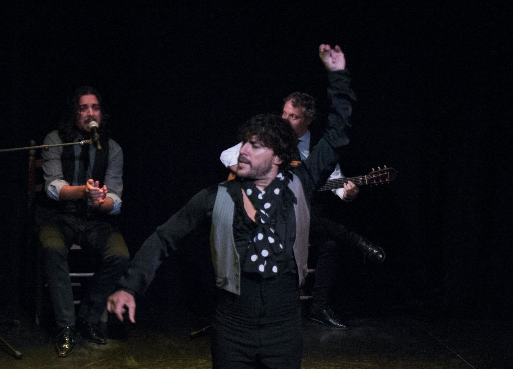 한 댄서가 두 팔을 들어 올린 채 공연하고, 뒤에 앉은 두 명의 뮤지션이 희미하게 빛나는 무대에서 노래를 부르고 기타를 연주합니다.