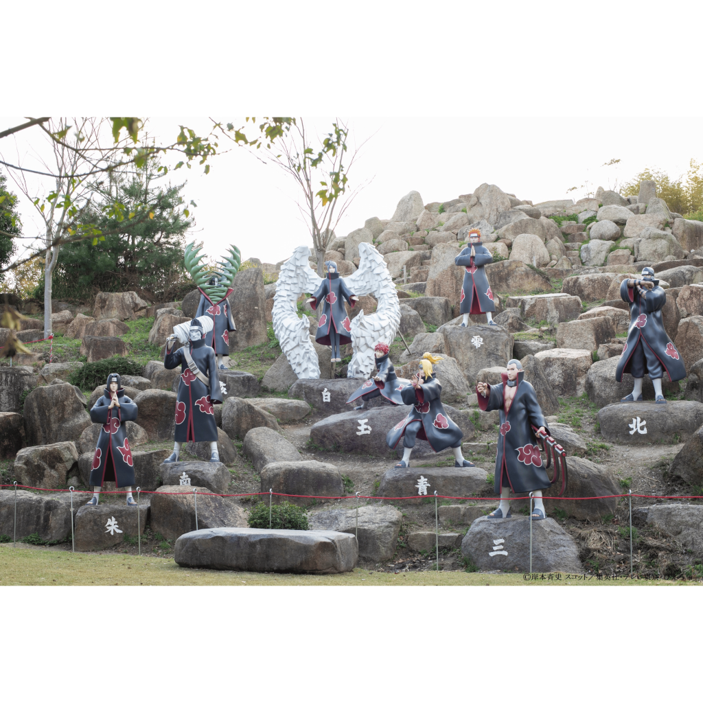 푸르름과 나무를 배경으로 바위 언덕에 배치된 검은 망토에 구름을 형상화한 캐릭터 조각상.