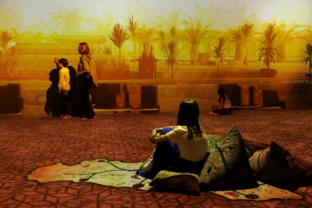 어린이와 성인은 야자수가 있는 사막 풍경이 펼쳐지는 몰입감 넘치는 전시물과 상호작용합니다. 어떤 사람들은 쿠션에 앉아 있습니다.