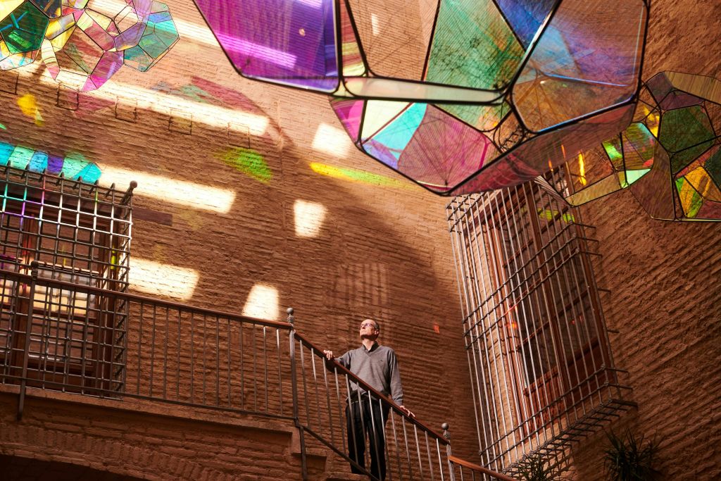 한 사람이 화려한 반투명 기하학적 패널 아래 계단에 서서 벽돌 벽에 가벼운 패턴을 시전하고 있습니다.