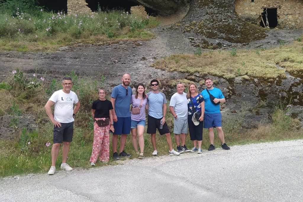 초목과 폐허를 배경으로 산비탈 근처의 도로에 7명의 사람들이 밖에서 서 있습니다.