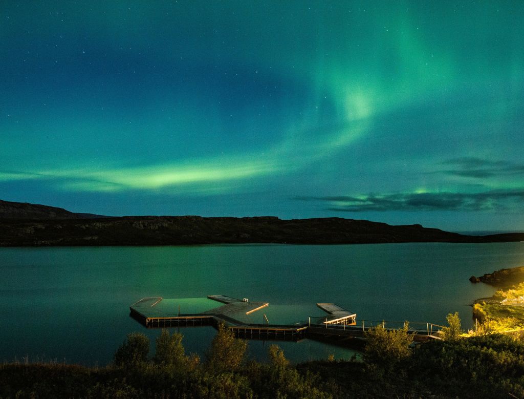 밤에는 고요한 호수에 나무로 된 선착장이 물로 뻗어 있고 오로라가 하늘을 비추고 있습니다.