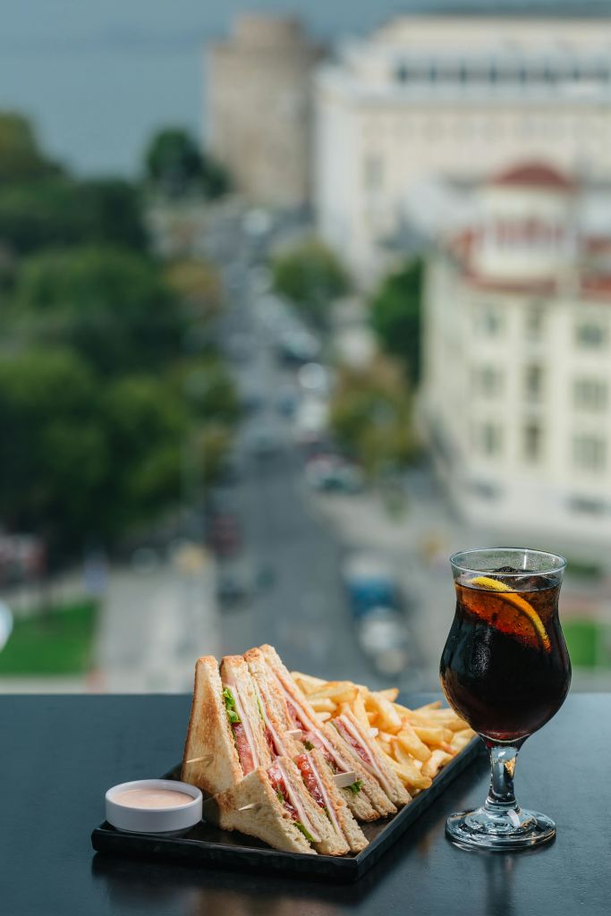 클럽 샌드위치, 감자튀김, 레몬이 든 탄산음료 한 잔이 테이블 위에 놓여 있으며, 도시 거리와 건물을 배경으로 내려다보입니다.