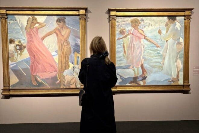 어두운 코트를 입고 흐르는 의복과 야외 장면의 인물이 그려진 두 개의 액자 그림을 관찰하는 사람.